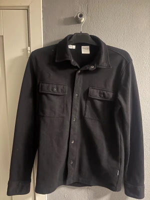Svart overshirt från Selected Homme - Svart overshirt från Selected Homme med klassisk krage och två stora bröstfickor med knappar. Storlek Small. Använd ett fåtal gånger så i väldigt fina skick. Pris kan diskuteras vid snabb affär. Tveka inte på höra av dig om du har funderingar. 