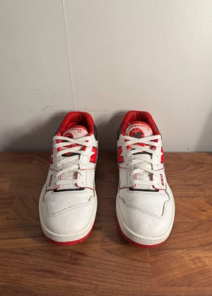 New Balance 550 skor - Säljer ett par New Balance 550 sneakers i vitt med röda och svarta detaljer. Skorna har snörning, platt sula och rund tå. Ovandelen är i skinn och textil, med tydlig New Balance-logga på sidan och 550-tryck. Perfekt för dig som gillar retrostil och sportig vibe.