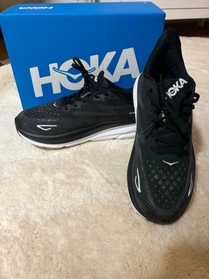 Svarta Hoka Clifton 9 sneakers - Säljer ett par svarta Hoka Clifton 9 sneakers med vit och svart sula. Skorna har mesh-överdel för bra ventilation, snörning och rund tå. Perfekta för löpning och träning, med lätt och stötdämpande sula som ger grym komfort. Hoka-logga på plösen och sidan.