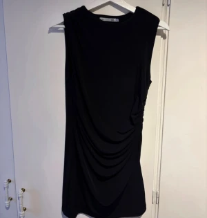 Svart omlott kortklänning från 157 - Svart ärmlös kortklänning från 157 i omlottstil med snygga draperingar på sidan. Klänningen har en enkel, stilren design och är gjord i mjukt syntetmaterial som sitter bekvämt mot kroppen. Köpt det här året och använt en gång. Storlek XS