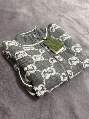 Gucci tröja  - Säljer en grå stickad kofta från Gucci med vit GG-mönster över hela plagget. Koftan har ribbad krage, muddar och nederkant, samt dragkedja framtill. Tillverkad i 100% ull och har två fickor med vit kant. Perfekt för dig som gillar exklusiv streetstyle.