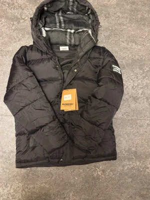Svart pufferjacka från Burberry - Svart pufferjacka från Burberry med huva och klassiskt rutigt foder i grått och vitt. Jackan har dragkedja och knappar framtill, samt Burberry London England-logga på ärmen. Perfekt för kalla dagar och har en riktigt schysst streetstil.