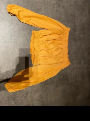 Gul offshoulder croppad blus - Säljer en snygg gul blus med offshoulder och långa ärmar. Toppen är croppad med smock vid axlar och midja, vilket ger en skön och trendig passform. Perfekt till jeans eller kjol för en färgstark look. Materialet känns lätt och luftigt.