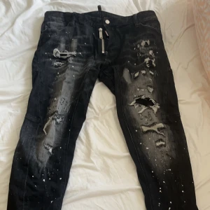 Svarta jeans från Dsquared2 - Säljer ett par svarta jeans från Dsquared2 med slitna detaljer och färgstänk. Jeansen har en dragkedja och knappar framtill samt en klassisk femficksdesign. Perfekta för en edgy look!