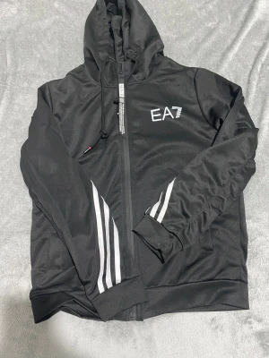 Svarta EA7 mjukisbyxor med vita ränder - Säljer ett par svarta mjukisbyxor från EA7 med vita ränder längs benen och logga på vänster sida. Byxorna har elastisk midja med snörning och dragkedjor vid fickorna med detaljerade band. Materialet är mjukt och syntetiskt, perfekt för chill eller träning.