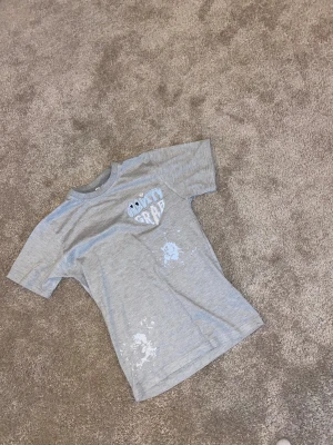 Grå Gravity Grab t-shirt med tryck - Snygg grå t-shirt från Gravity Grab med broderad text och ögon på bröstet. T-shirten har coolt vitt färgstänk-tryck framtill och är i mjuk bomull. Klassisk passform och korta ärmar, perfekt för en chill och avslappnad stil.