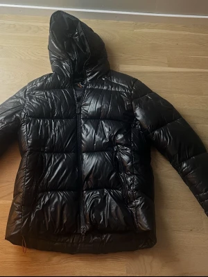 Svart pufferjacka ifrån Save The Duck - Svart pufferjacka från Save The Duck med glansig finish och huva. Jackan har två sidofickor med dragkedja och tydlig '100% Animal Free'-text på insidan. Perfekt för kalla dagar. Inga defekter. Storlek S men passar även M.
