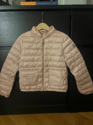 Beige Moncler pufferjacka - Säljer en beige/rosa pufferjacka från Moncler med klassisk logga på ärmen. Jackan har två fickor med knappar framtill, hög krage med knapp och dragkedja. Den är quiltad och har en snygg, stilren look som funkar till många outfits. Jackan är i ett väldigt bra skick och fåtal gånger använd! 