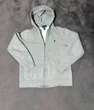 Grå hoodie från Polo Ralph Lauren - Snygg grå hoodie från Polo Ralph Lauren med dragkedja och huva. Tröjan har långa ärmar, ribbade muddar och en klassisk Polo-logga broderad på bröstet. Perfekt för vintern och enkel att matcha med jeans❄️ Storlek L(14-16). Sitter som XS