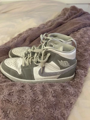 Nike Air Jordan 1 Highs grå/vita - Säljer ett par Nike Air Jordan 1 Highs i grått och vitt. Skorna har klassisk snörning, perforerad tå och rund tå. Swoosh-loggan på sidan och Air Jordan-vingen på ankeln. Materialet är en mix av skinn och syntet. Perfekt för dig som gillar streetwear och sneakers med ikonisk design.