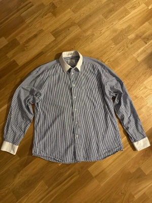 Randig skjorta från Baccante  - Snygg blåvit randig skjorta från Ralph Lauren med vit krage och vita muddar. Klassisk passform, långärmad och med knappar framtill. Skjortan har en bröstficka och är gjord i mjuk bomull som känns skön mot huden. Perfekt för dig som gillar stilrena detaljer.