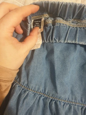 Blå långkjol med volanger från SHEIN - Säljer en blå långkjol från SHEIN i storlek 12-13 år. Kjolen har flera volanger och ett elastiskt midjeband för skön passform. Materialet är mjukt och lätt, perfekt för dig som gillar denimlooken men vill ha något bekvämt och luftigt.