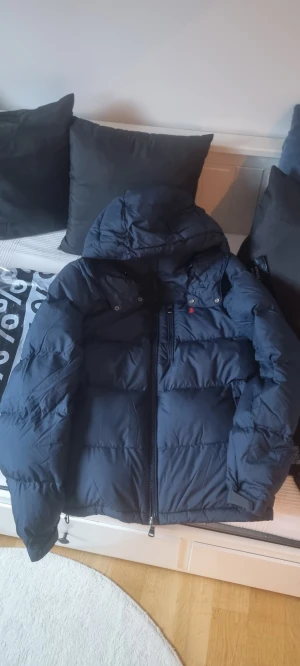 Mörkblå pufferjacka från Polo Ralph Lauren - Snygg mörkblå pufferjacka från Polo Ralph Lauren med huva och flera praktiska fickor med dragkedja. Jackan har ett broderat rött Polo-logo på bröstet och är tillverkad i polyester. Perfekt för kalla dagar och har en normal passform.