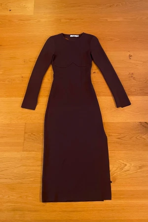 Mörkbrun långärmad långklänning från NAKD - Snygg mörkbrun långklänning från NAKD med långa ärmar och figurnära passform. Klänningen är öppen i ryggen och har en slits vid vänster ben. Den går lite under knäna och är i stretchigt material. Lite stor i storleken.
