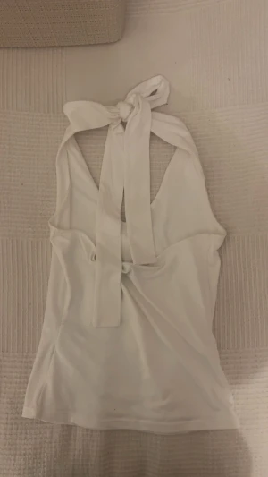 Vit topp med knytband och rosett - Säljer en stilren vit topp med breda axelband som knyts i nacken till en rosett. Toppen har en öppen rygg och är ärmlös, perfekt för varma dagar. Köpt utomlands och i storlek 8, vilket motsvarar runt XS/XXS. Skriv för fler bilder eller frågor💗