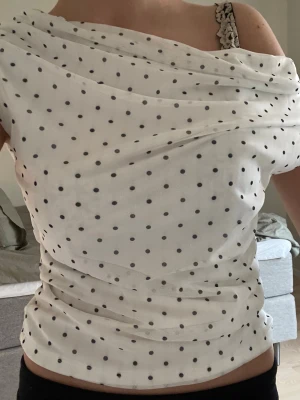 Prickig offshoulder topp Gina Tricot - Säljer en vit offshoulder topp från Gina Tricot med svarta prickar. Toppen har ett omlottliknande tyg som ger en snygg draperad effekt och är ärmlös. Materialet känns lätt och luftigt, perfekt för varmare dagar. Supersnygg att styla med jeans eller kjol!
