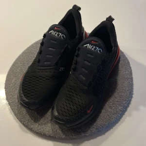 Nike Air Max 270 svarta/röda sneakers storlek 40 - Nike Air Max 270 sneakers i svart med röda detaljer och stor synlig Air-enhet i hälen. Ovandelen är i ventilerande syntetmaterial med diskret snörning och små vita loggor. Snygg och sportig look som sticker ut med den röda sulan och de röda swoosh-loggorna. (Har inte snören)