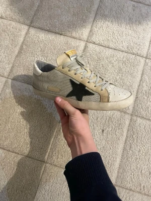 Golden goose skor  - Säljer ett par snygga sneakers från Golden goose Sneakers med beige och vita paneler, grå snörning och en svart stjärna på sidan. Ovandelen är i textil och skinn, med perforerad tå och gummisula. Perfekt för dig som gillar stilrena detaljer och cool design.