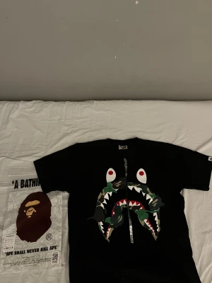 Svart BAPE Shark t-shirt med camo - En äkta Svart t-shirt från BAPE i storlek S med ikonisk Shark-motiv på bröstet och detaljer i camo. Klassisk rund hals och korta ärmar. BAPE-logga på ärmen och originalförpackning medföljer. Materialet är mjuk bomull som känns skönt mot huden. Den är köpt från GOAT