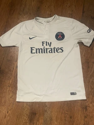 PSG fotbollströja storlek L - Bilden visar en vit Paris Saint-Germain fotbollströja från Nike med Fly Emirates tryck, inte en sko. Ingen skorelaterad information kan ges.