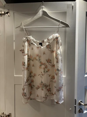 Blommig blus från Vero Moda - Säljer en söt blommig blus från Vero Moda i storlek M. Blusen är vit med tunna axelband och har ett romantiskt blommönster i rosa och grönt. Den har volangdetaljer vid halsringningen och är tillverkad i ett lätt och luftigt syntetmaterial.