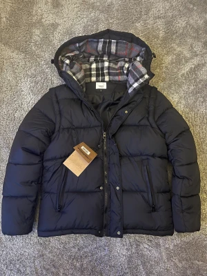 Svart pufferjacka från Burberry - Svart pufferjacka från Burberry med avtagbara ärmar, så den kan användas som väst. Jackan har huva med klassiskt rutigt Burberry-foder i grått, vitt och rött. Två dragkedjefickor fram och logotyp på ena ärmen. Perfekt för kalla dagar.