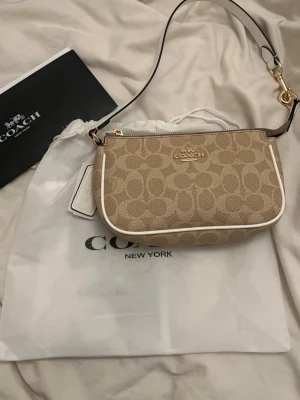 Beige axelväska  - Säljer en snygg beige axelväska från Coach  Nolita 19 med klassiskt monogrammönster och vita detaljer. Väskan har guldfärgade metalldetaljer, dragkedja och en vit axelrem. Från en affordable luxury plattform. Du får med dustbag. Nypris 900kr