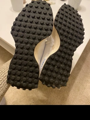 New Balance sneakers med blå detalj - Säljer ett par fräscha New Balance sneakers i vitt med beige mockadetaljer och en stor blå logga på sidan. Skorna har grov, svart gummisula med rundade pluppar och snörning framtill. Perfekt för dig som gillar sportig stil och snygga färgkombinationer.