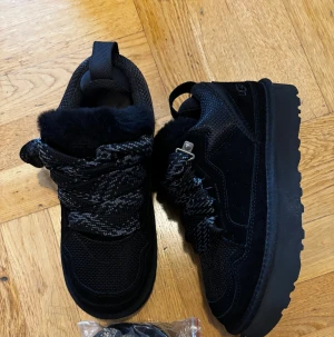 UGG LOWMEL - Säljer ett par svarta UGG sneakers med fluffigt pälsfoder och grova snören i svart och grått. Skorna har platt sula med tydligt mönster undertill och detaljer i mocka och syntet. Perfekta för dig som vill ha både stil och komfort under höst och vinter. STORLEKEN är 41 men sitter mindre mer som 39-40