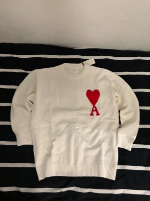Vit stickad tröja från Ami Paris - Vit stickad tröja från Ami Paris med rund hals och ribbade muddar. På bröstet finns ett stort rött hjärta och bokstaven A som broderad detalj. Tröjan har lång ärm och är gjord i mjukt material som känns skönt mot huden. Hör av dig om du har några frågor.