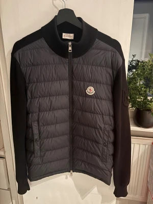 Svart quiltad pufferjacka från Moncler - Svart pufferjacka från Moncler med quiltad framsida och stickade ärmar och rygg. Jackan har ribbad krage, två fickor med knappstängning och dragkedja framtill. Moncler-logga på bröstet ger en clean och exklusiv vibe.