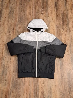 Svart och vit vindjacka med huva - Snygg vindjacka i svart, grått och vitt med huva och dragsko. Jackan har en rak passform, långa ärmar och praktiska fickor framtill. Materialet är syntet och polyester, perfekt för blåsiga dagar. Stängs med vit dragkedja hela vägen upp.