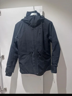 Svart vindjacka från Everest med huva - Svart vindjacka från Everest med huva och dragkedja framtill. Jackan är vattentät med TCS Waterproof-teknologi och har praktiska fickor med dragkedja. Storlek 164 