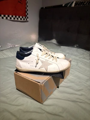 Golden Goose vita sneakers med stjärna - Pris kan diskuteras! Snygga vita sneakers från Golden Goose med grå stjärndetalj på sidan och mörkblå häl. Skorna har snörning och platt sula, tillverkade i skinn med beige inslag på sulan. Perfekta för dig som gillar en clean men ändå unik look.