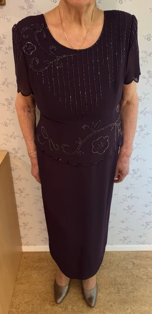 Mörklila långklänning R&M Richards - Elegant mörklila långklänning från R&M Richards by Karen Kwong, dekorerad med glittriga pärlor i blommönster och vertikala linjer. Klänningen har korta ärmar, rund halsringning och vågig kant nedtill. Materialet är 100% polyester och klänningen har dragkedja bak.