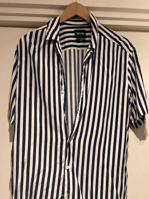 Randig skjorta från Only & Sons - Säljer en kortärmad skjorta från Only & Sons i marinblått och vitt med vertikala ränder. Skjortan har klassisk krage, knappar framtill och en relaxed fit. Tillverkad i mjuk bomull som känns skön mot huden. Perfekt för dig som gillar stilrena och fräscha plagg.