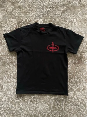 Svart Corteiz t-shirt  - Svart t-shirt från Slaughter Gang med fet röd print på ryggen och mindre tryck framtill. Snyggt svärdmotiv och texten 'Slaughter Gang' i stor stil. Klassisk passform och gjord i mjuk bomull. Perfekt för dig som gillar streetwear och vill sticka ut.