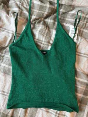 Grön glittrig linnetopp från H&M - Säljer en snygg grön linnetopp från H&M i storlek XS. Toppen har tunna axelband, v-ringning och är glittrig i materialet vilket ger en cool effekt. Perfekt jul eller festtopp att styla med jeans eller kjol för en trendig look.
