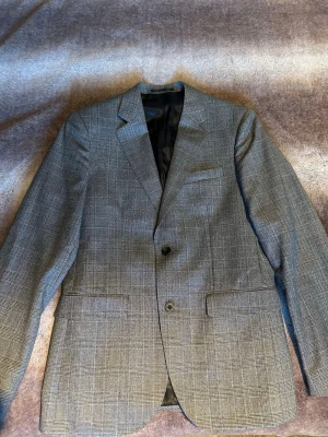 Grå rutig kavaj från Bläck - Snygg grå kavaj från Bläck med klassiskt rutig mönster och två knappar framtill. Skräddad passform och stilren design med fickor och slät insida. Perfekt för dig som vill ha en clean och modern look.