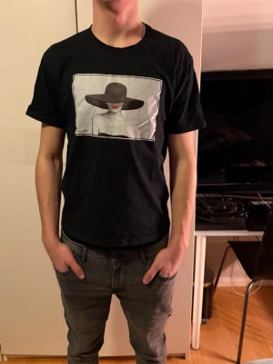 Svart t-shirt med tryck från CelemonAce - Svart t-shirt från CelemonAce i storlek M. Framsidan har ett stort tryck med en person i hatt i svartvitt. Klassisk passform med rund hals och korta ärmar. Skön och mjuk känsla, perfekt till jeans eller shorts.