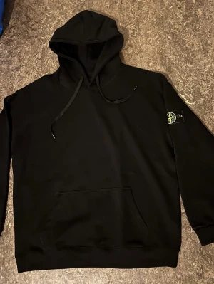 Stone Island-hoodie - Stone Island-hoodie i klassiskt svart utförande. Plagget är i fint skick med den ikoniska kompasslogotypen på ärmen. Perfekt för den som söker en stilren och tidlös tröja. 