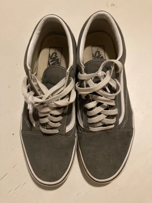 Grå Vans Old Skool sneakers - Säljer ett par grå Vans Old Skool sneakers med vit sula och klassisk vit sidostripes. Skorna har snörning och är tillverkade i en kombination av mocka och canvas. Yttersulan är i gummi med det typiska rutmönstret. Perfekt för dig som gillar streetstyle och skate.