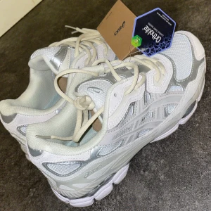 Vita och silver Asics GEL sneakers - Fräscha Asics GEL sneakers i vit och silver med mesh och syntetpaneler. Skorna har snörning, rund tå och platt sula. Perfekt för dig som gillar sportig stil och vill ha bekväma skor med snygga detaljer. Asics-logga på sidan och GEL-detalj bak.