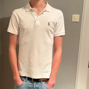 Vit kortärmad pikétröja från Ralph Lauren - Säljer en klassisk vit pikétröja från Ralph Lauren med korta ärmar och en broderad logotyp på bröstet. Perfekt för en stilren och avslappnad look.