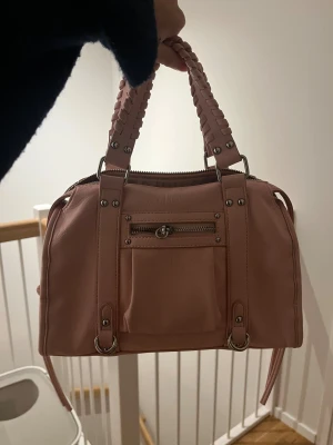 Rosa handväska från Amor med dragkedja - Säljer en ljusrosa handväska från Amor med snygga flätade handtag och silverfärgade detaljer. Väskan har ett praktiskt ytterfack med dragkedja och rymligt innerfack. Materialet är mjukt syntet och designen är stilren med metallnitar och ringar.