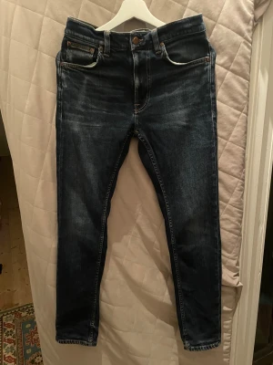 Mörkblå jeans från Nudie Jeans - Säljer ett par mörkblå jeans från Nudie Jeans i storlek 30/30. Modellen har klassisk kontrastsömmar och snygga slitningar/wash för en snygg archive look. Jeansen är tillverkade i bomull och har en slim passform med raka ben. Lean Dean är modellen!