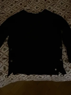 Svart stickad tröja från Zink med knappar - Säljer en svart stickad tröja från Zink i storlek S. Tröjan har lång ärm och coola silverfärgade knappar längs sidorna som ger en snygg detalj. Perfekt för dig som gillar stilrena och enkla plagg med en twist.