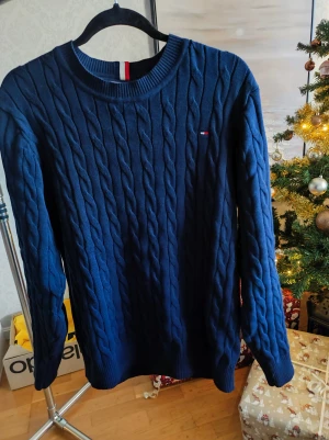 Marinblå stickad tröja Tommy Hilfiger - Säljer en marinblå stickad tröja från Tommy Hilfiger i storlek L. Tröjan har ett klassiskt kabelstickat mönster, rund halsringning och långa ärmar. Diskret logga broderad på bröstet och ribbade muddar vid hals, ärmslut och nederkant. Perfekt för kyliga dagar.