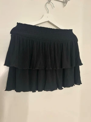 Svart volangkjol från Kappahl - Säljer en svart kortkjol från Kappahl med dubbla volanger och smock i midjan. Kjolen har en luftig och flowy känsla, perfekt för dig som gillar en enkel men snygg look. Materialet är lätt och bekvämt, och den svarta färgen gör den enkel att matcha med olika toppar. Utstrechad på bilden💗