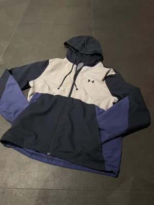 Under Armour vindjacka med huva - Snygg colorblock vindjacka från Under Armour i mörkblått, grått och blått. Jackan har huva med snörning, dragkedja framtill och meshfoder. Perfekt för dig som vill ha en sportig look och gillar funktionella detaljer. Den är riktigt skön och är en perfekt vår jacka som håller värme bra men också går att ha på sommaren. Priset är inte hugget i sten. 